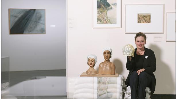 Eine Frau sitzt in einer Galerie neben zwei Büsten und hält eine Badekappe hoch.