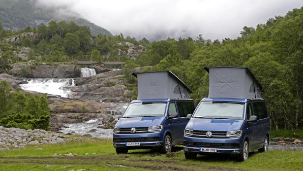 Zwei blaue VW-Campingbusse stehen vor einer malerischen Flusslandschaft mit Wasserfall.