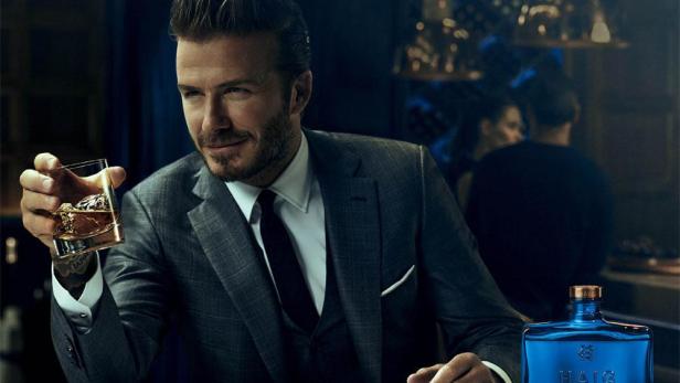 David Beckham in einem grauen Anzug hält ein Glas Whisky in einer Bar.