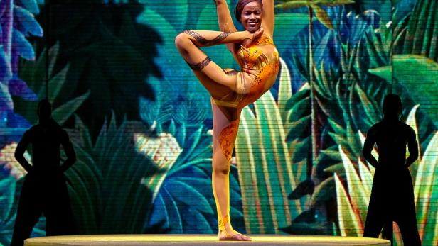 Eine Akrobatin in einem orangefarbenen Kostüm steht auf einem Bein auf einer Bühne vor einem Dschungelhintergrund.