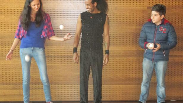 Eine Frau, ein Mann und ein Junge jonglieren mit weißen Bällen vor einer Holzwand.