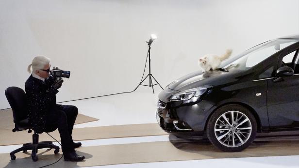Karl Lagerfeld fotografiert eine Katze, die auf der Motorhaube eines schwarzen Autos sitzt.