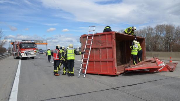 Ein umgekippter roter Container liegt auf einer Autobahn, umgeben von Feuerwehrleuten.