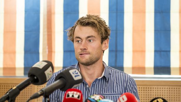 Ein Mann mit blonden Haaren steht vor Mikrofonen.