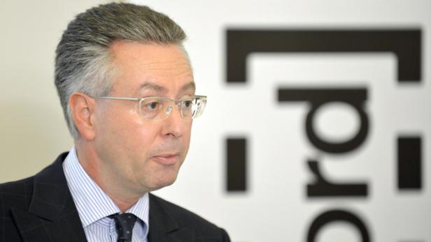 Ein Mann mit Brille und Anzug vor einem unscharfen Logo.