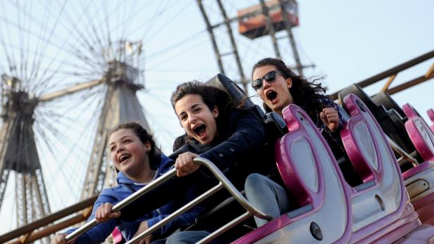 Drei junge Frauen schreien aufgeregt in einer Achterbahn vor einem Riesenrad.