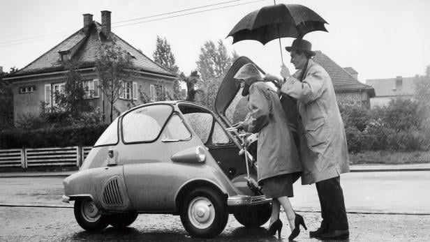 Ein Mann hält einen Regenschirm über eine Frau, die in ein Isetta-ähnliches Auto einsteigt.