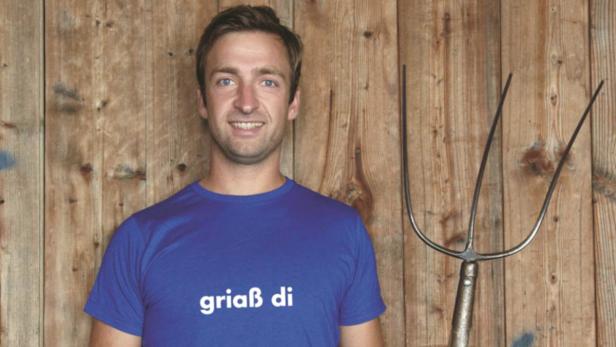 Ein Mann mit einem blauen T-Shirt mit der Aufschrift „griaß di“ und einer Mistgabel vor einer Holzwand.