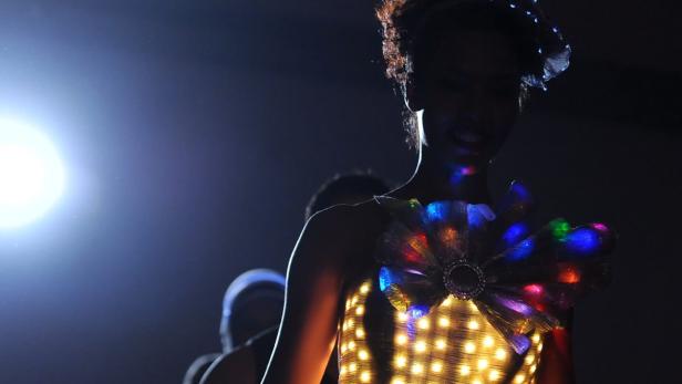 Eine Person trägt ein leuchtendes Kleid mit bunten Lichtern auf einer dunklen Bühne.