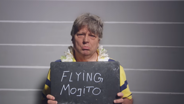 Ein Mann mit Blumenkette hält ein Schild mit der Aufschrift „Flying Mojito“ vor einer Fotowand.