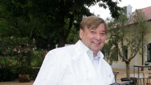 Ein lächelnder Mann in Kochkleidung steht im Freien vor einem Gebäude.