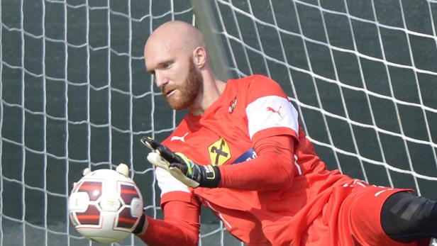 Ein Torwart in roter Sportkleidung fängt einen Fußball vor einem Tornetz.