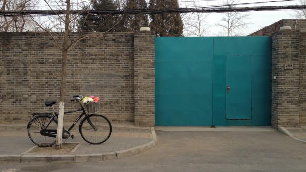 Ein Fahrrad mit Blumenkorb steht vor einer türkisfarbenen Tür in einer Backsteinmauer.