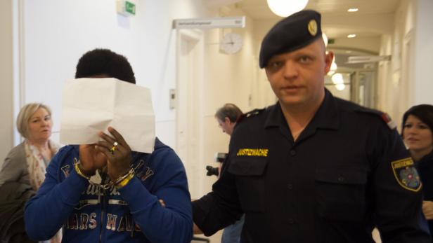 Ein Gefangener mit Handschellen hält ein Blatt Papier vor sein Gesicht, während ein Justizbeamter daneben steht.