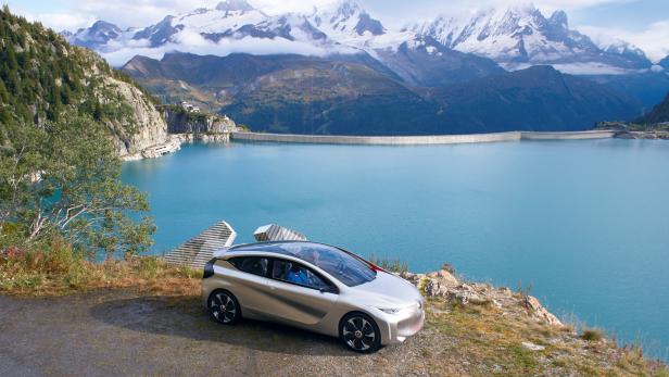Ein silbernes Auto steht vor einem Bergsee mit einem Staudamm im Hintergrund.