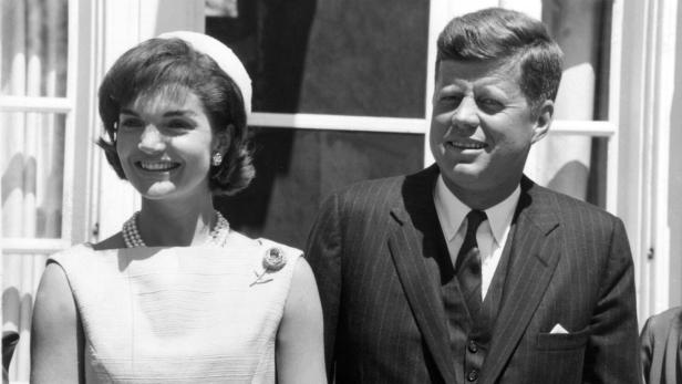 John F. Kennedy und Jacqueline Kennedy lächeln nebeneinander stehend vor einem hellen Gebäude.