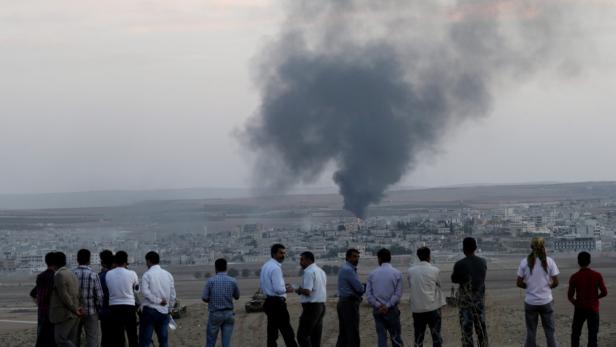 Eine Gruppe von Männern beobachtet eine Rauchsäule, die über einer Stadt aufsteigt.