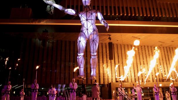 Eine riesige, beleuchtete Roboter-Skulptur steht vor einem Gebäude, umgeben von Fackelträgern.