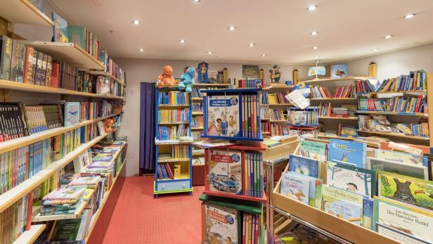 Regale voller bunter Kinderbücher, einige Stofftiere sitzen auf den oberen Regalen, helle Beleuchtung und roter Boden.