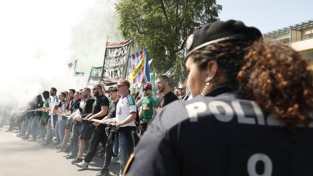 Eine Polizistin steht vor einer Gruppe Fußballfans mit Fahnen und Rauch.
