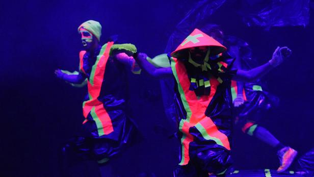 Zwei Personen in leuchtenden, futuristischen Kostümen treten im Dunkeln auf.