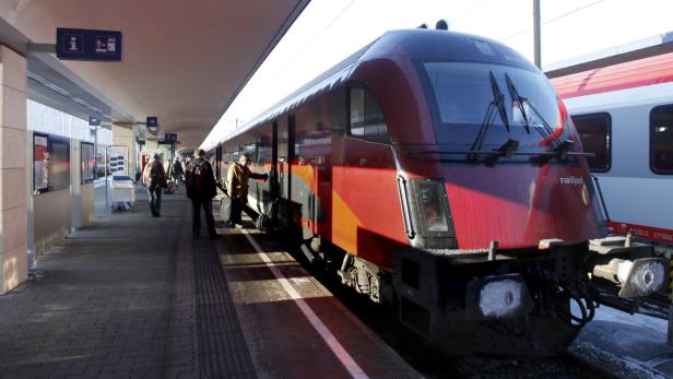 Ein roter Railjet-Zug steht an einem Bahnsteig mit wartenden Reisenden.
