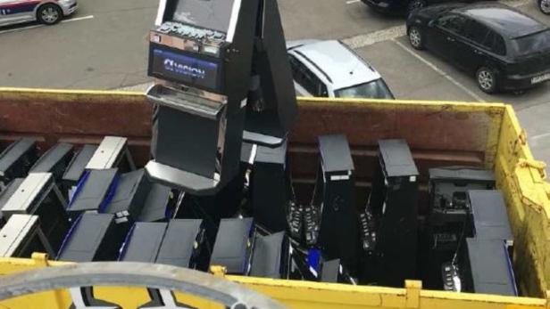 Ein Schrottgreifer hebt einen Spielautomaten über einen gelben Container voller weiterer Automaten.