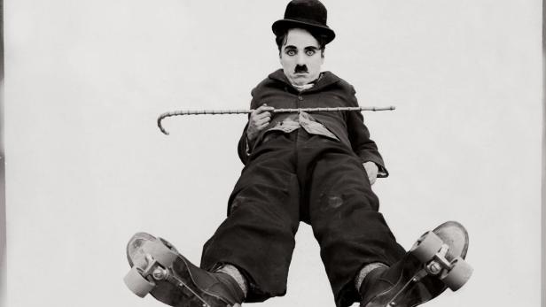 Charlie Chaplin sitzt auf Rollschuhen und hält einen Stock in der Hand.