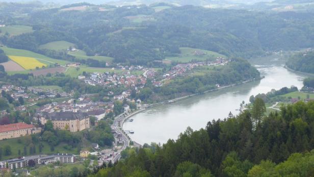 Blick auf eine Stadt mit einem Schloss an einem Fluss, umgeben von bewaldeten Hügeln.