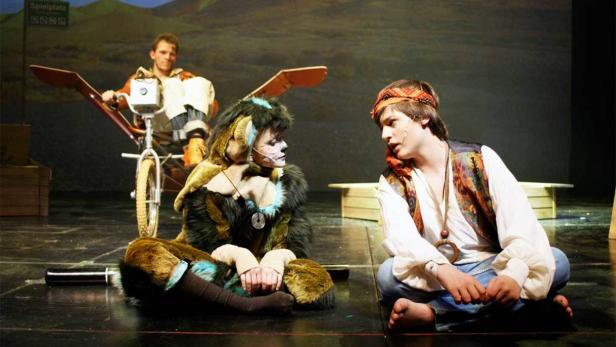 Zwei Schauspieler in Kostümen sitzen auf einer Bühne, während ein dritter auf einem Fahrrad im Hintergrund steht.