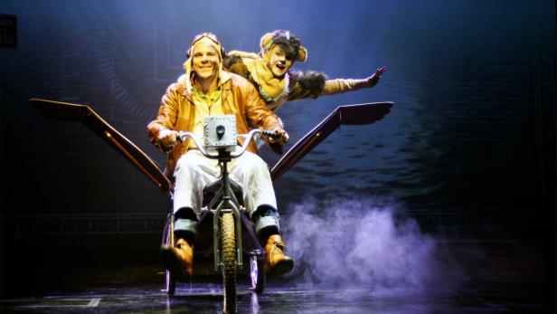 Ein Mann mit Fliegerbrille fährt ein seltsames Flugrad, während ein Mann im Katzenkostüm hinter ihm steht.