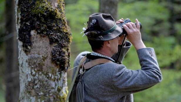 Ein Mann mit Trachtenhut beobachtet mit einem Fernglas die Umgebung im Wald.