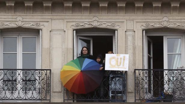 Eine Frau und ein Kind stehen auf einem Balkon mit einem Regenbogen-Regenschirm und einem Schild mit der Aufschrift „Oui“.