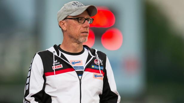 Ein Mann mit Brille und Kappe, bekleidet mit einer Jacke mit Sponsorenlogos.