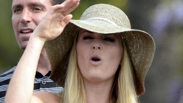 Eine blonde Frau mit Hut grüßt mit erhobener Hand, ein Mann steht im Hintergrund.