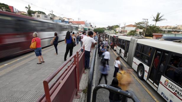 Menschen warten an einer Bushaltestelle in São Paulo, während Busse vorbeifahren.