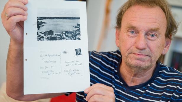 Ein Mann hält ein Blatt Papier mit einer Schwarzweißfotografie einer Stadt am Wasser in der Hand.