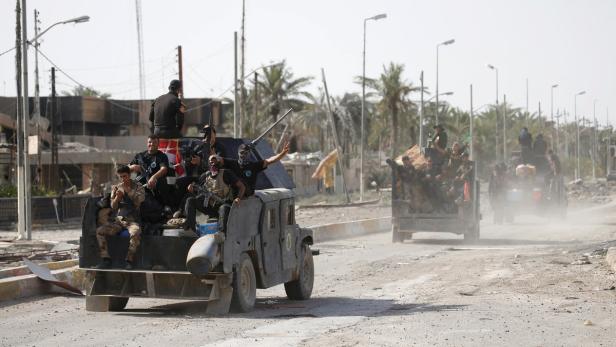 Bewaffnete Männer fahren auf Militärfahrzeugen durch eine zerstörte Stadt.