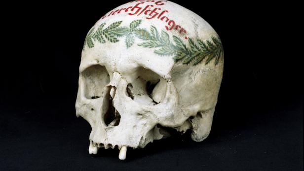 Ein menschlicher Schädel mit aufgemalter Inschrift und einem Lorbeerkranz.