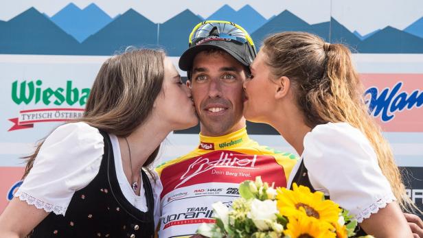 Ein Radrennfahrer wird von zwei Frauen auf dem Siegerpodest geküsst.