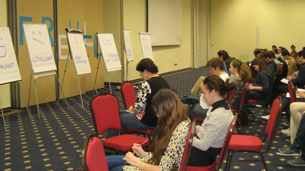 Zuschauer sitzen in einem Raum vor Flipcharts mit Aufschriften wie „Highlight“ und „LOL“.