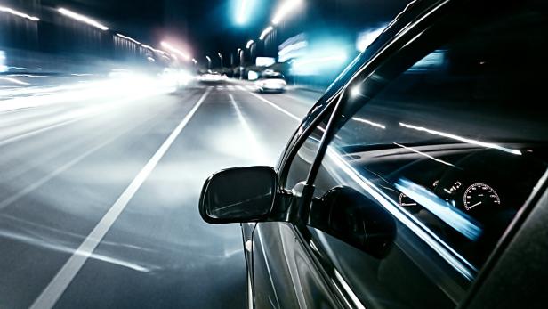 Ein Auto fährt nachts mit hoher Geschwindigkeit auf einer beleuchteten Straße, Lichter ziehen Spuren.