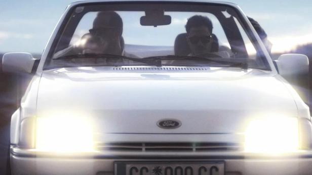 Ein weißer Ford Escort mit zwei Personen auf einer Landstraße.