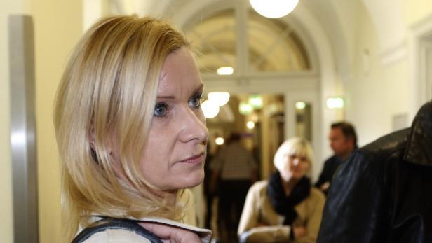 Eine blonde Frau mit ernstem Blick steht in einem belebten Flur.