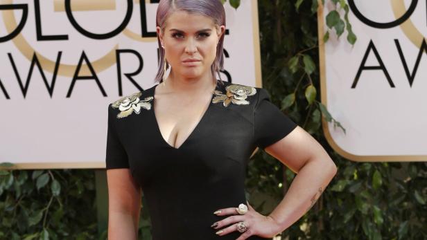 Kelly Osbourne posiert auf dem roten Teppich in einem schwarzen Kleid mit goldenen Applikationen.