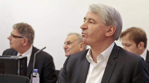 Ein Mann mit grauem Haar und Anzug sitzt bei einer Konferenz in der ersten Reihe.