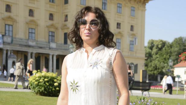 Eine Frau mit runder Sonnenbrille steht vor einem gelben Gebäude.