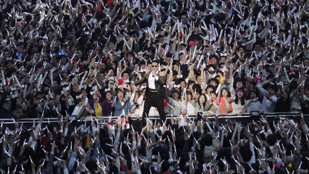 PSY tritt vor einer jubelnden Menschenmenge mit Leuchtstäben auf.