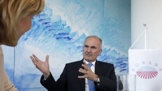 Ein Mann im Anzug gestikuliert während eines Gesprächs vor einem blauen Hintergrund und einer Flagge.