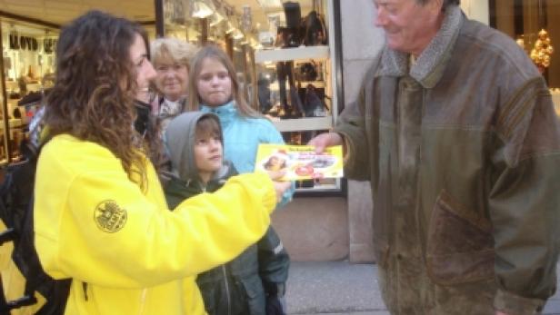 Eine Frau in gelber Jacke übergibt einem Mann eine Broschüre, während Kinder zusehen.
